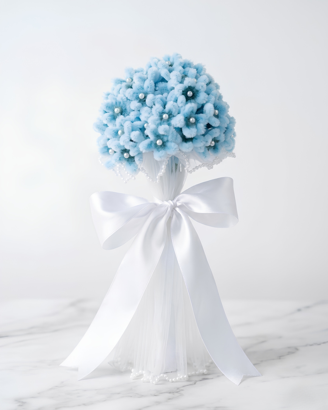 Hortensia celeste eterna hecha a mano en chenille, con perlas entre los pétalos, velo de tul y gran lazo de raso blanco, sobre mármol blanco