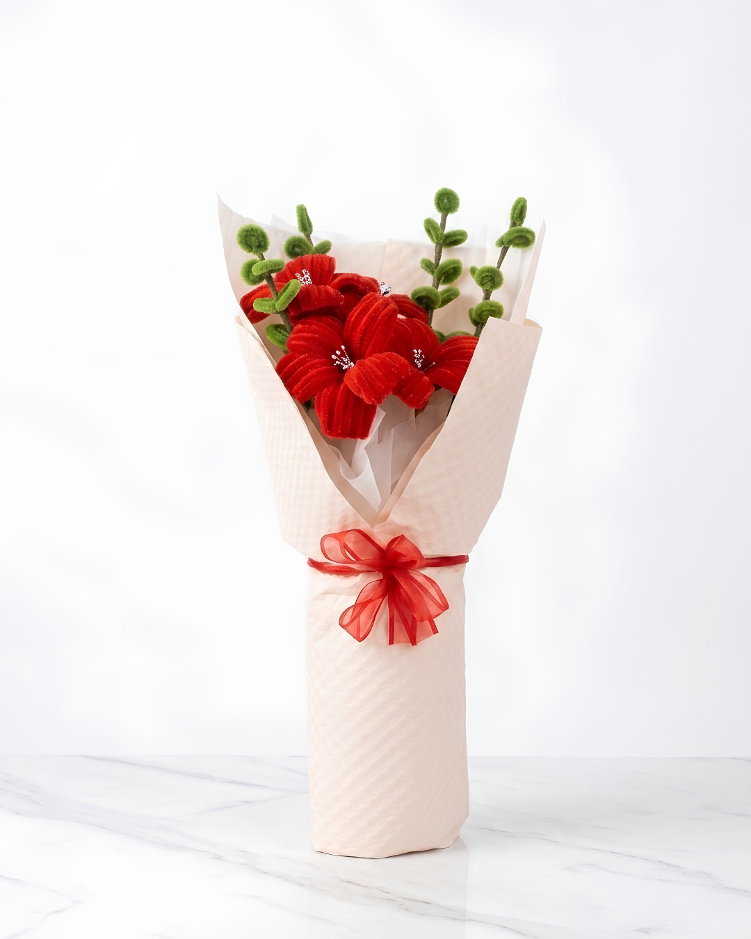 Ramo de flores rojas eternas hechas a mano en chenille, con hojas verdes, envuelto en papel rosa palo con lazo de organza roja, sobre mármol blanco