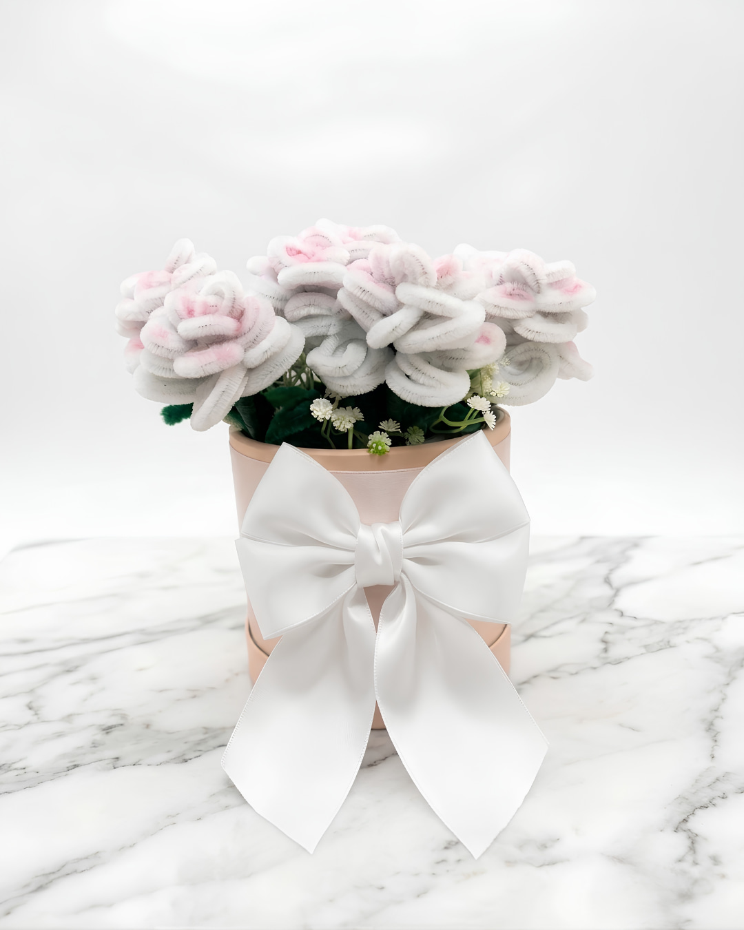 Arreglo de flores blancas con reflejos rosados hechas a mano en chenille, con gypsophila, en caja redonda color nude con gran lazo de raso blanco, sobre mármol blanco