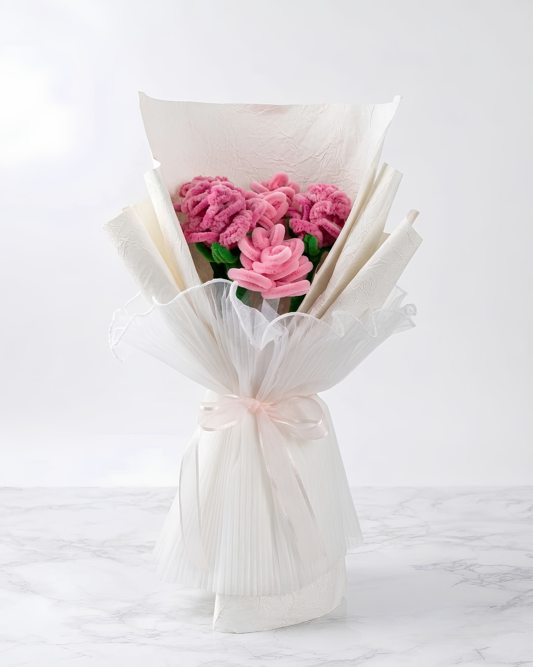 Ramo de rosas rosadas eternas hechas a mano en chenille, envuelto en papel blanco plisado y tul con lazo de cinta rosa, sobre mármol blanco